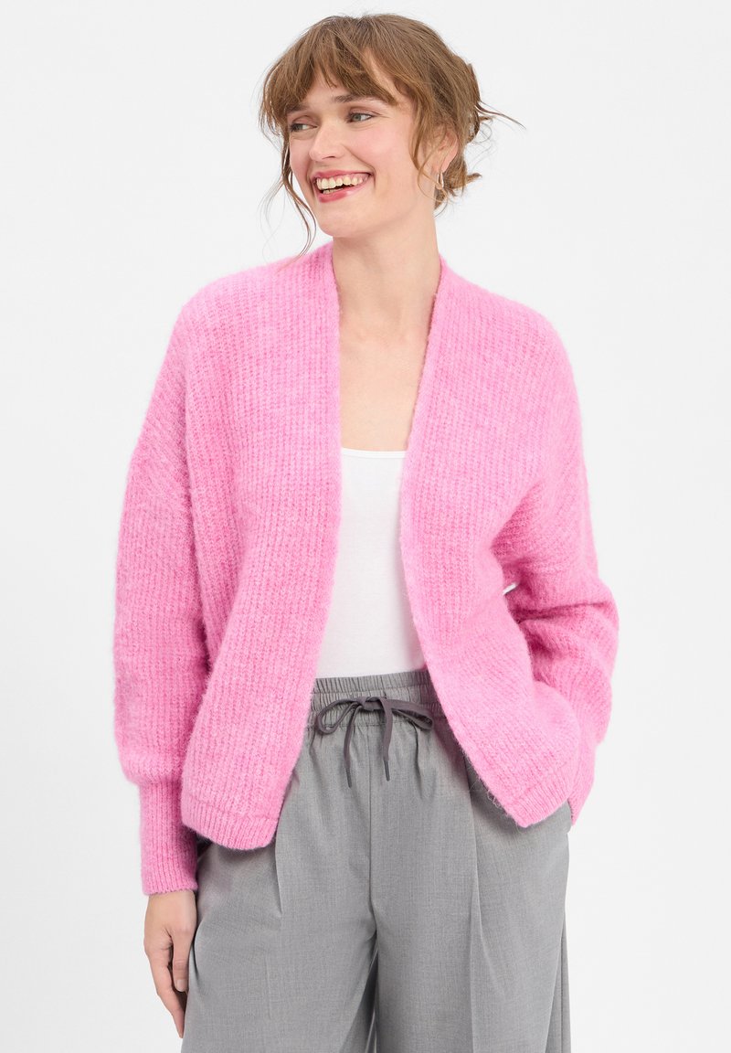 Rosa gestrickte Strickjacke mit einem lockeren, offenen Design, langen Ärmeln und strukturierter Ripp-Optik, kombiniert mit hellgrauen Sweatpants mit Kordelzug.