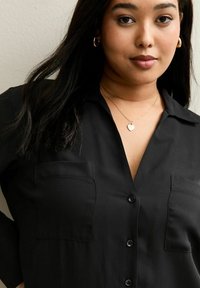 Młoda kobieta z długimi ciemnymi włosami nosi złote kolczyki koła, naszyjnik z zawieszką w kształcie serca oraz czarną koszulę na guziki z dwoma kieszeniami na piersi.