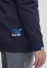 Crna dukserica s rebrastom teksturom, sa plavim izvezenim logotipom "!SLD" i "Od 1993." na prednjoj strani i rukavu.