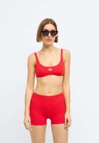 Γυναίκα που φοράει κόκκινο crop top και ταιριαστά σορτς με ψηλή μέση, με κοντά μαλλιά και μεγάλα μαύρα γυαλιά ηλίου, στέκεται μπροστά σε ένα απλό φόντο.