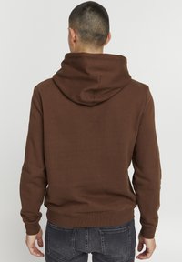 Brun hættejakke med snor i hætten og ribbede manchetter. Blødt stof med en ensartet overflade. Vist på en person, der vender væk fra kameraet.