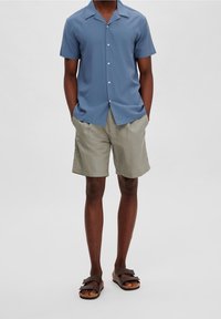 Ljusblå kortärmad skjorta med notch-krage, i kombination med beiga shorts och bruna sandaler med platt sula och justerbara remmar.