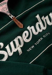 Groene sweatshirt met een kwart-rits hals, voorzien van grote witte geborduurde tekst "Superdry" en "New York City," met een leren rits trekker in bruin.