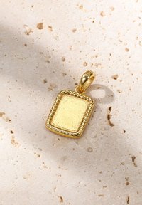 Pendentif en or de forme carrée avec une surface texturée, présentant un bord décoratif en corde. Placé sur un fond en pierre neutre.