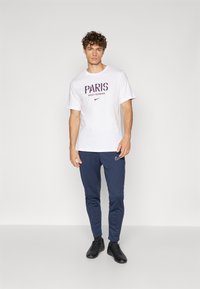 Balta medvilninė marškinė su "PARIS SAINT-GERMAIN" grafika mėlyna ir raudona spalvomis. Suderinta su tamsiai mėlynomis sportinėmis kelnėmis ir juodais sportiniais batais.