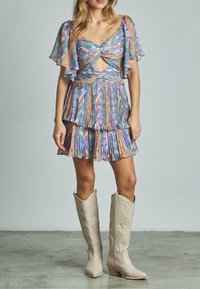 Farbenfrohes, plissiertes Minikleid mit Flatterärmeln, einer Twist-Front und einem geschichteten Rock. Trägt kniehohe beigefarbene Cowboystiefel.