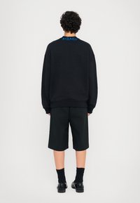 Sweatshirt preto oversized com bordado azul "MILANO" nas costas, combinado com calções pretos e sapatos pretos, visto por trás.