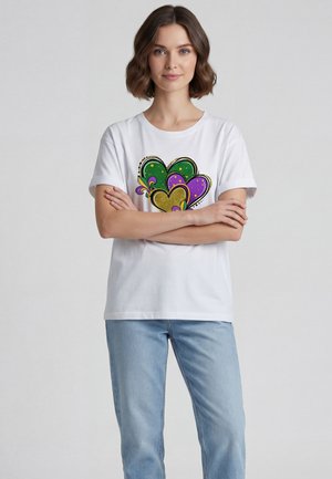 Femme aux cheveux courts bruns portant un t-shirt blanc avec des motifs de cœurs colorés et un jean bleu clair, debout les bras croisés.