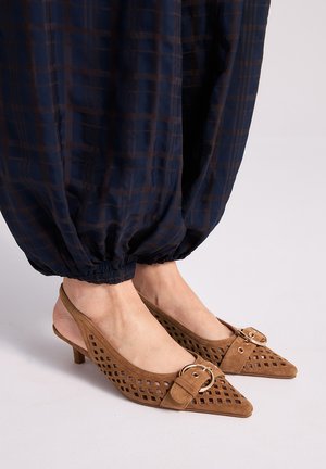 Pies con sandalias de tacón bajo tipo kitten slingback de ante marrón con detalles recortados y hebillas doradas, combinadas con pantalones oscuros a cuadros con puños elásticos.