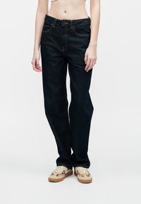 Oseba, oblečena v temne ravne hlače in bež superge z �živalskim potiskom, stoji pred enotno belo ozadjem.