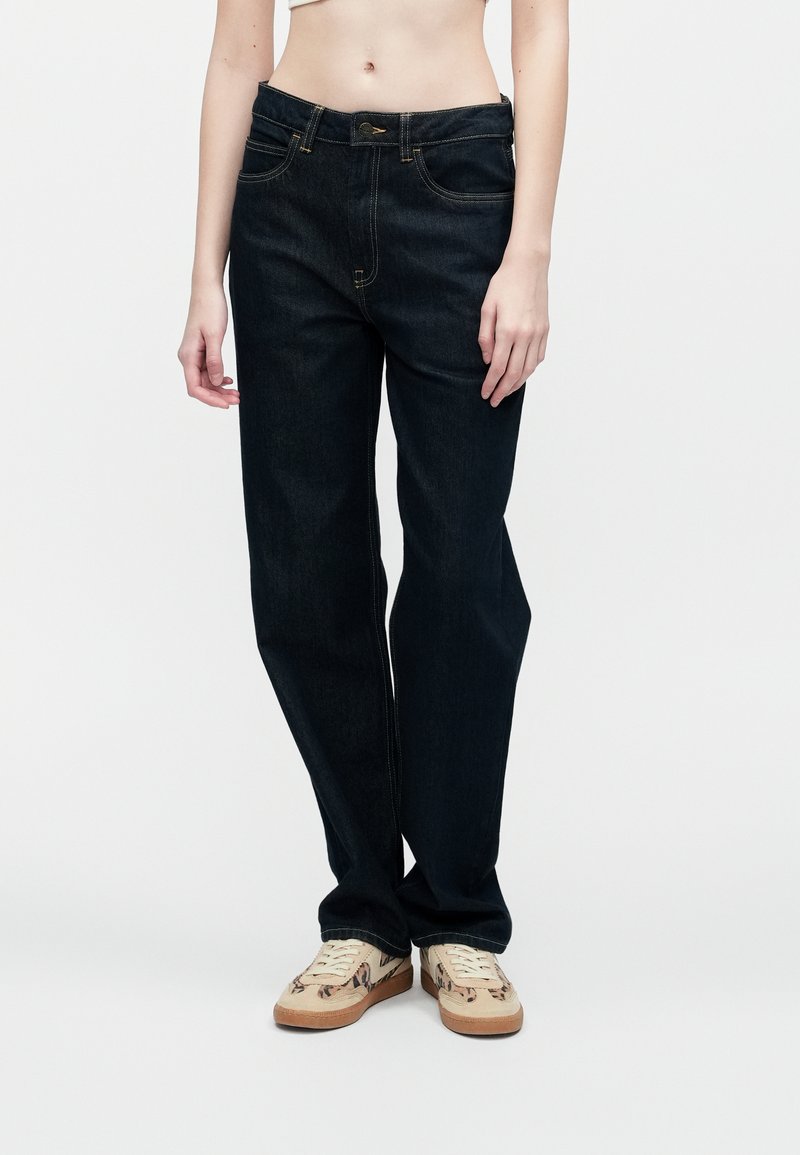 Oseba, oblečena v temne ravne hlače in bež superge z živalskim potiskom, stoji pred enotno belo ozadjem.