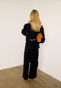 Γυναίκα με μακριά ξανθά μαλλιά στέκεται με την πλάτη σε λευκό τοίχο, φοράει μαύρο ρούχο με πουά σχέδιο και καφέ πέδιλα με τακούνι, κρατώντας ένα καφέ πορτοφόλι.