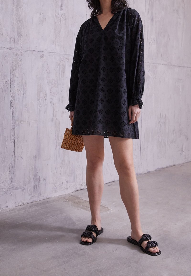 Kvinde iført en sort minikjole med lange ærmer og hulmønstre, der holder en lille brun flettet håndtaske og har sorte flettede slid-sandaler på.