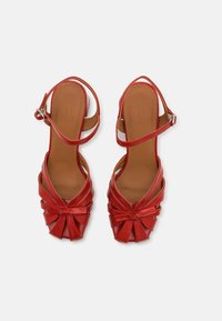 Punase nahkasaapad kõrge kontsaga, keerduva esiosa kujundusega, avatud varvastega ja ankle strap'iga. Sile tekstuur koos pehmendatud beež sisetallaga.