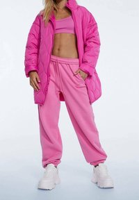 Puffermantel und Sweatpants-Set in leuchtendem Pink. Der Mantel hat eine gesteppte Textur, einen Reißverschluss und große Taschen. Die Sneaker sind weiß und klobig.