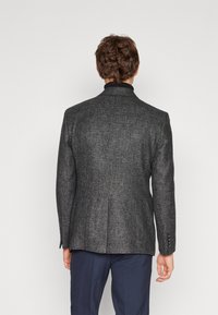 Blazer a medida en un gris oscuro con tejido texturizado, abertura simple en la parte trasera y tres botones en el puño. Combinado con pantalones azul marino.