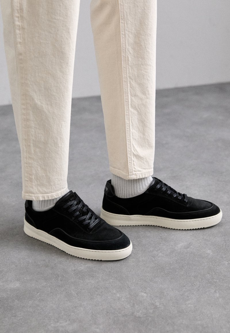 Black suede sneakers with a white rubber sole, round toe, and textured detailing. Paired with light beige pants and grey crew socks.