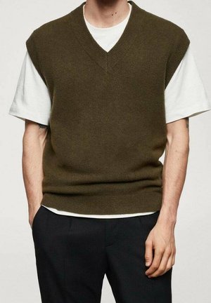 Person iført en olivengrøn strikket ærmeløs sweater over en hvid t-shirt med korte ærmer og sorte bukser, med hænderne i lommerne.