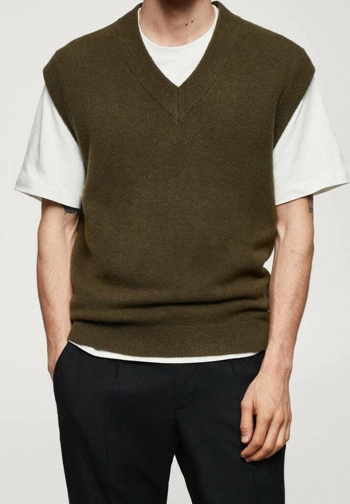 Personne portant un pull sans manches en maille vert olive par-dessus un t-shirt blanc à manches courtes et un pantalon noir, les mains dans les poches.