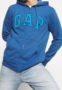 Sweat à capuche zippé bleu avec "GAP" en grandes lettres porté par une personne posant les mains derrière le dos et les jambes écartées, portant un jean bleu clair.