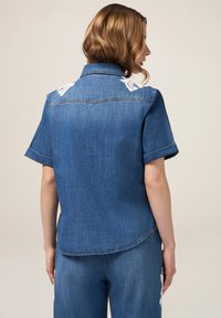 Camicia di jeans di blu medio, maniche corte, con accenti ricamati bianchi sulle cuciture delle spalle, orlo curvo e cuciture visibili.