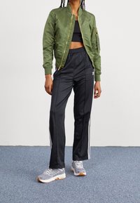Veste bomber en satin vert avec fermeture éclair dorée, crop top noir, pantalon de sport noir avec bandes blanches, et chaussures de sport grises à semelle gomme.