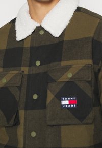 Rutig skjortjacka i mörkgrön och svart flanell, med en vit fårskinnskrage, gröna metalltryckknappar och två bröstfickor med logotyp.