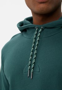 Grüne Kapuzenjacke aus weichem Stoff, mit einem Kordelzug mit geflochtenem Muster und Metallspitzen sowie einem dezenten Logo auf der Brust.