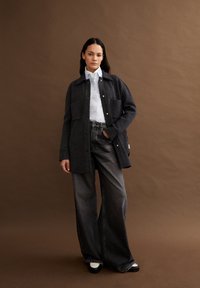 Giacca oversize in lana grigia con due tasche frontali, indossata sopra una camicia bianca con bottoni e jeans in denim scuro a gamba larga. Scarpe in bianco e nero.