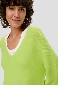 Sweter w kolorze limonkowej zieleni z dekoltem w kształcie litery V, z fakturowanym wzorem i kontrastującą białą podszewką kołnierza. Miękki, lekki materiał.