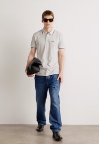 Polo cinza claro com detalhes em preto, jeans de ganga azuis e óculos de sol pretos. Asegurando uma carteira preta, de pé sobre um fundo neutro.