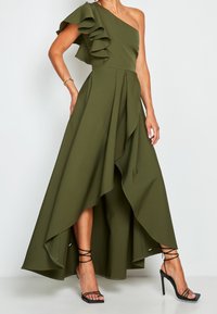 Robe olive verte, à une épaule, avec une manche volante, ourlet asymétrique et design superposé. Associée à des escarpins à talons hauts noirs à bretelles.