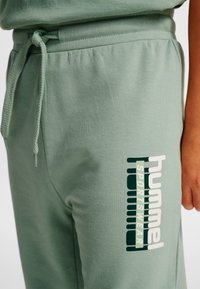 Close-up van lichtgroene joggingbroek met trekkoord in de taille en "hummel SPORTSWEAR"-logo op de dij, gedragen door een persoon.