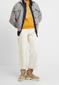 Rutig grå och svart ulljacka över en orange sweatshirt med logotyp, tillsammans med vita cropped byxor och beiga vandringsskor.