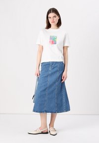 T-shirt blanc à manches courtes avec un imprimé graphique coloré, associé à une longue jupe en denim bleu. Modèle portant des chaussures claires.