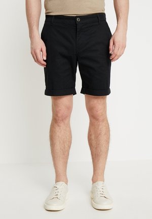 Mann trägt schwarze, umgekrempelte Shorts und weiße Turnschuhe, steht vor einem schlichten weißen Hintergrund.