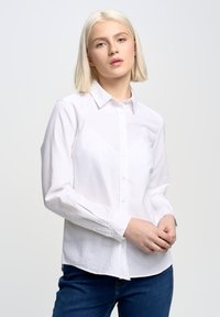 Camisa blanca de algodón con botones, con cuello, mangas largas y un corte ajustado, combinada con jeans de color azul oscuro. Textura suave y diseño clásico.