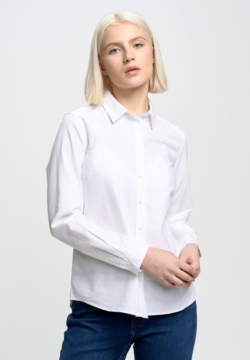 Camisa blanca de algodón con botones, con cuello, mangas largas y un corte ajustado, combinada con jeans de color azul oscuro. Textura suave y diseño clásico.
