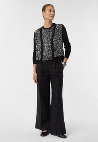 Schwarze und beige Strickjacke mit Leopardenmuster, langen Ärmeln und Knöpfen, kombiniert mit lockeren schwarzen Hosen und schwarzen Schuhen. Einfaches Design.