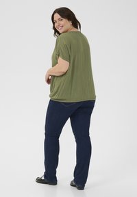 Blusa holgada de color verde oliva, de mangas cortas y con dobladillo redondeado, combinada con jeans azul oscuro y zapatos negros. Un look sencillo y casual.