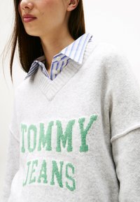 Grauer Pullover mit V-Ausschnitt und grüner "TOMMY JEANS"-Aufschrift. Über einem blau-weiß gestreiften Hemd mit Kragen getragen. Weicher Stoff, lässiges Design.