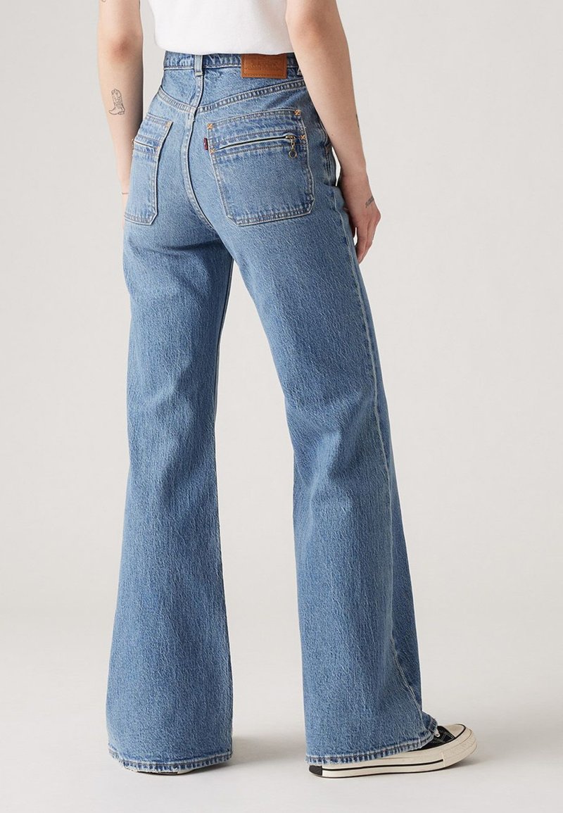 Jeans évasés en denim bleu délavé avec une taille haute, comportant deux poches arrière et une étiquette en cuir au niveau de la taille. La texture est légèrement usée.
