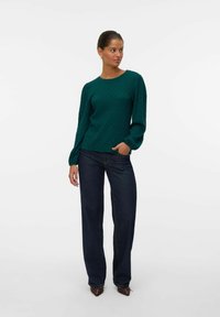 Teal strukturerad tröja med långa puffärmar, i kombination med mörkblå raka jeans och spetsiga skor, stående mot en vit bakgrund.