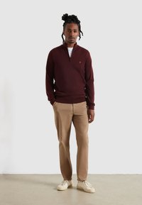 Maglione bordeaux con zip e texture a coste, abbinato a pantaloni beige e sneakers bianche sporche, in piedi davanti a uno sfondo neutro.