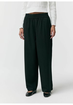 Kobieta ubrana w luźne czarne spodnie w prążki, biały krótki sweter dzianinowy oraz czarne płaskie buty, stojąca na tle białej ściany.