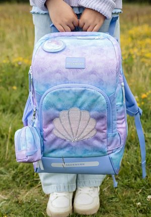 Bambino che tiene uno zaino pastello viola e blu con motivo a conchiglia, in piedi sull'erba con scarpe da ginnastica bianche visibili.