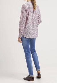 Chemise à manches longues de couleur violet clair avec un motif circulaire, associée à un jean bleu et des chaussures marron. Présente un détail boutonné dans le dos.