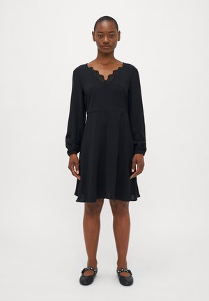 Black dress with long sleeves, V-neckline featuring a scalloped lace trim, flared hem, and made of a textured fabric. Paired with black flats.