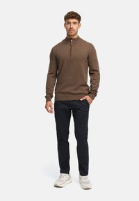 Brun zip-up tröja med texturerad stickning, med hög krage och ribbade muddar, kombinerad med mörkblå byxor och ljusa sneakers.