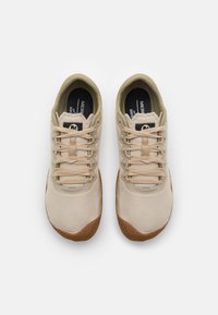 Beige canvas-sneakers med beige snören, gummiyttersula och texturerat grepp. Skorna har rundad tå och svarta insidesmärken som kontrast.